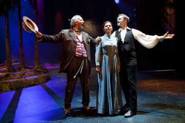 Lavalla seisoo kolme aikakauden pukuihin pukeutunutta näyttelijää; yksi mies huutaa iloisesti hattua kohottaen, sinipukuinen nainen seisoo hänen ja valkoiseen paitaan pukeutuneen toisen miehen välissä, molemmat kädet ojennettuina, dramaattisessa teatterikohtauksessa.