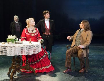 Neljä näyttelijää esiintyy lavalla aikakauden puvuissa; punamekkoon pukeutunut nainen istuu pöydän ääressä raidalliseen pukuun pukeutuneen miehen kanssa, kun toinen smokkiin pukeutunut mies seisoo heidän välissään, ja taustalla seisoo vanhempi mies.