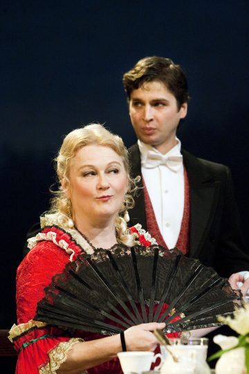 Koristeelliseen punaiseen mekkoon pukeutunut nainen pitelee mustaa viuhkaa ja katsoo sivulle, kun taas hänen takanaan tummalla taustalla seisoo muodolliseen pukuun, rusettisolmioon ja punaisiin liiveihin pukeutunut mies.