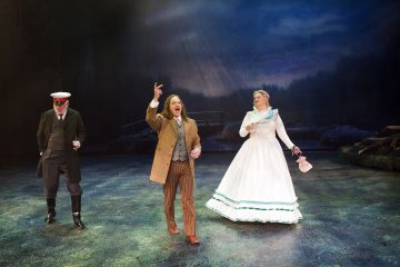 Lavalla esiintyy kolme näyttelijää: yksi puvussa ja lippiksessä katselee alaspäin, toinen ruskeassa takissa ja raidallisissa housuissa elehtii tarmokkaasti ja kolmas valkoisessa aikalaispuvussa viuhka kädessään metsätaustaa vasten.