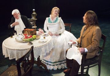 Kolme aikakauden pukuihin pukeutunutta näyttelijää istuu pöydän ympärillä, jossa on pitsinen pöytäliina, hedelmiä ja teesetti, ja näyttävät nauttivan vilkkaasta keskustelusta näyttämöllä teatteriesityksen aikana.