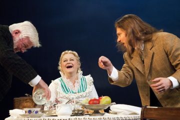 Kolme aikakauden pukuihin pukeutunutta näyttelijää esiintyy pöydän ääressä, jossa on ruokaa ja astioita. Keskellä oleva nainen nauraa, ja kaksi miestä molemmin puolin elehtii eloisasti vilkkaan näyttämökohtauksen aikana.