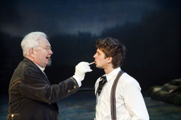 Vanhempi mies, jolla on silmälasit ja valkoiset hanskat, osoittaa kepillä tai työkalulla nuoremman miehen suuta, jolla on paita ja housunkannattimet, tummalla, epäselvällä taustalla.