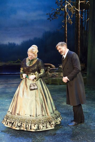 Pitkään, koristeelliseen vihreään mekkoon pukeutunut nainen seisoo lavalla mustaan pukuun pukeutuneen miehen vieressä, ja molemmat hymyilevät hieman metsäaiheisessa teatterikuvassa.