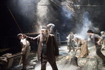 Näyttelijät esiintyvät näyttämöllä dramaattisessa valaistuksessa, ja yksi mies on edessä pitelemässä sauvaa ja muut hänen takanaan lakaisevat lehtiä. Kohtaus on dynaaminen, ja siinä on liikettä ja savua, mikä tuo mieleen historiallisen tai teatterin tunnelman.