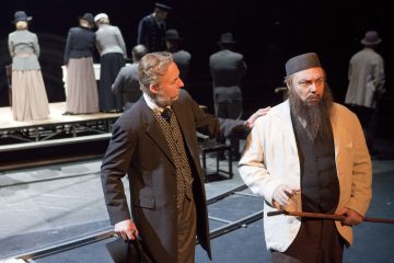 Kaksi aikakauden pukuihin pukeutunutta miestä seisoo näyttämön etualalla, toisella on parta ja keppi, toinen koskettaa olkapäätään. Taustalla korotetulla lavalla seisoo useita vanhanaikaisiin vaatteisiin pukeutuneita ihmisiä.