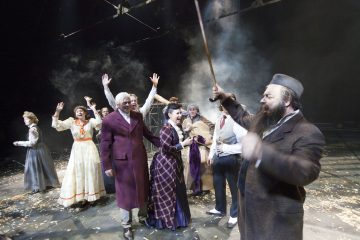 Lavalla esiintyy joukko aikakauden pukuihin pukeutuneita näyttelijöitä; yksi mies edessä elehtii kepillä, kun taas toiset hänen takanaan, jotkut kädet kohotettuna, reagoivat dramaattisesti savuisessa valaistuksessa.