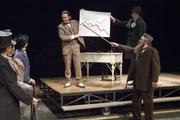 Lavalla esiintyy viisi vintage-pukuihin pukeutunutta näyttelijää. Yhdellä on kädessään kaavio, jossa on punaisia ja mustia viivoja, ja toinen osoittaa sitä tikulla. He seisovat pöydän ympärillä ja näyttävät keskustelevan taulukosta vilkkaasti.