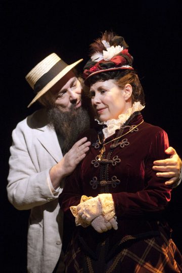 Vaaleaan pukuun ja olkihattuun pukeutunut mies syleilee viktoriaaniseen tyyliin pukeutunutta naista, jolla on viktoriaaninen mekko ja hattu. Miehellä on parta ja hän näyttää puhuvan naiselle, joka näyttää mietteliäältä ja hieman tunteelliselta.