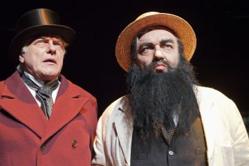 Kaksi pukumiestä seisoo vierekkäin. Toisella on tumma silinterihattu, ruskeankeltainen takki ja harmaa solmio; toisella on olkihattu, valkoinen takki, raidallinen paita ja suuri musta tekoparta. Molemmat katsovat vakavin ilmein eteenpäin.