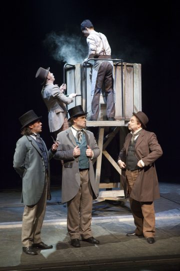 Kolme 1800-luvun asuun ja silinterihattuihin pukeutunutta miestä seisoo lavalla puhumassa, kun kaksi muuta työskentelee savun kanssa heidän yläpuolellaan olevan puurakenteen takana teatteriesityksessä.