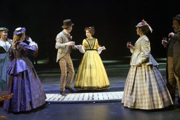 Lavalla esiintyy useita viktoriaanisen ajan pukuihin pukeutuneita näyttelijöitä, ja keskellä on yksi keltaiseen mekkoon pukeutunut nainen. Muut värikkäisiin pukuihin ja pukuihin pukeutuneet pitävät laseja kädessään ja näyttävät käyvän vilkasta keskustelua.