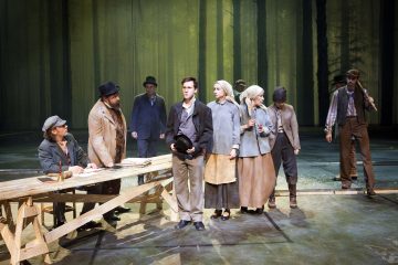 Ryhmä aikakauden pukuihin pukeutuneita näyttelijöitä esiintyy näyttämöllä korkeiden puiden muodostaman taustan edessä. Jotkut puhuvat, kun taas toiset seisovat jonossa puupöydän vieressä, mikä viittaa dramaattiseen tai historialliseen kohtaukseen.