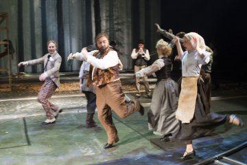 Ryhmä aikakauden pukuihin pukeutuneita näyttelijöitä tanssii energisesti lavalla, taustalla soittavat muusikot ja heidän takanaan on metsäaiheinen lavastus. Kohtaus vangitsee vilkkaan liikkeen ja juhlatunnelman.