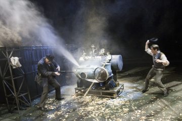 Kaksi näyttelijää on näyttämöllä vuorovaikutuksessa suuren, steampunk-tyylisen, savua tai höyryä päästävän koneen kanssa, kun lehdet peittävät maan ja dramaattinen valaistus lisää kohtauksen voimakkuutta.