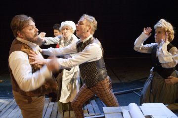 Liiviin pukeutunut mies työntää toista miestä rinnasta dramaattisen näyttämöteatteriesityksen aikana, kun kolme muuta näyttelijää aikakauden puvuissa reagoi huolestuneena ja yllättyneenä.