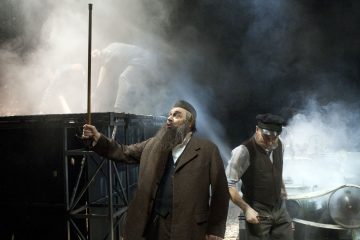 Parrakas mies ruskeassa takissa ja hatussa pitelee keppiä ja elehtii intohimoisesti savuisella näyttämöllä, kun toinen lippalakkipäinen mies katsoo alaspäin ja taustalla työskentelee useita hahmoja.