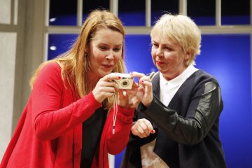 Kaksi naista seisoo lähekkäin sisätiloissa, hymyilee ja katsoo digitaalikameraan. Toinen punapukuinen nainen pitelee kameraa, kun taas toinen mustavalkoiseen asuun pukeutunut nainen katsoo näyttöä. Taustalla on ikkuna, jossa on sininen valo.