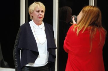 Lyhyet vaaleat hiukset omaava nainen seisoo kameraan päin, kun toinen punaisiin pukeutunut, pitkät punaiset hiukset omaava henkilö ottaa hänestä kuvan sisätiloissa.