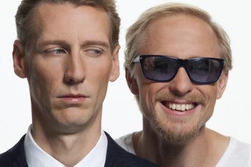 Kaksi miestä seisoo lähekkäin; vasemmalla oleva mies näyttää vakavalta ja vilkaisee sivulle, kun taas oikealla oleva mies hymyilee leveästi ja käyttää aurinkolaseja. He ovat valkoista taustaa vasten.