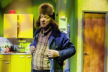 Vanhempi mies, jolla on karvainen talvihattu ja sininen takki, seisoo kirkkaasti valaistussa keittiössä ja näyttää mietteliäältä. Taustalla näkyy keltaisia kaappeja ja suljettu puuovi.