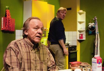 Kaksi miestä on värikkäässä keittiössä; toinen istuu aamiaistavaroita sisältävän pöydän ääressä ja näyttää huolestuneelta, kun taas toinen seisoo lieden vieressä ja vilkuilee taakseen. Kohtaus vaikuttaa jännittyneeltä tai dramaattiselta.