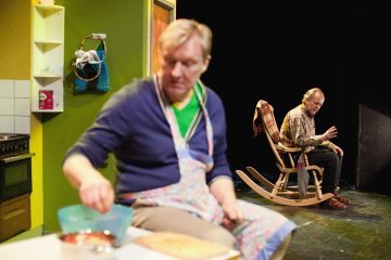 Kaksi miestä on näyttämöllä sisällä; toinen istuu puisessa keinutuolissa heiluttaen, kun taas toinen, esiliina päällään, on etualalla ja valmistelee ruokaa keittiön tiskillä.