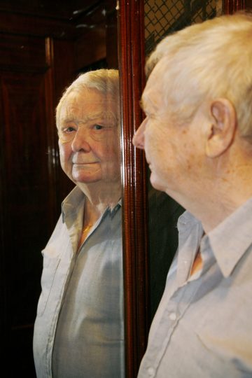 Iäkäs, valkotukkainen ja vakavalla ilmeellä varustettu mies seisoo peilin edessä, ja hänen peilikuvansa näkyy hänen vieressään. Hänellä on yllään vaaleansininen paita hämärässä huoneessa, jonka seinät ovat puiset.