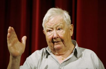 Iäkäs harmaahiuksinen mies osoittaa eleillä vasenta kättään kohottaen samalla kun hän puhuu. Hänellä on vaaleanharmaa paita ja kultainen kaulakoru. Taustalla näkyy punainen verho.