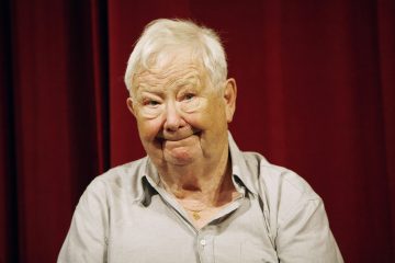 Valkohiuksinen ja vaaleanharmaapaitainen iäkäs mies hymyilee lämpimästi punaisen verhotaustan edessä.