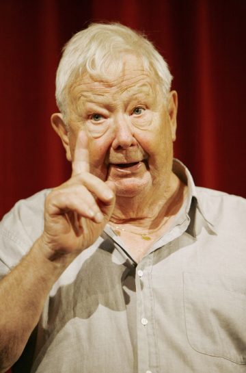 Iäkäs mies, jolla on lyhyet valkoiset hiukset ja vaaleanharmaa paita, elehtii nostamalla oikean etusormensa ylös. Hän seisoo syvän punaisen verhon edessä ja näyttää puhuvan tai esittävän jotain.