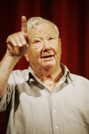 Iäkäs harmaahiuksinen mies, jolla on vaalea paita, osoittaa oikealla kädellään ylöspäin sormella. Hän seisoo punaisen verhon edessä ja näyttää puhuvan tai puhuvan yleisölle.