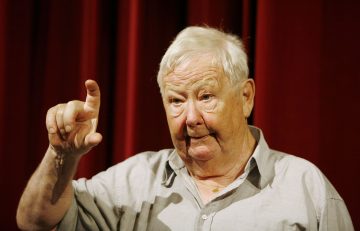 Valkohiuksinen iäkäs mies, jolla on vaaleanharmaa paita, osoittaa oikealla kädellään ylöspäin. Hän seisoo punaisen verhon edessä ja hänen ilmeensä on ilmeikäs.