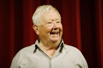 Valkohiuksinen iäkäs mies hymyilee lämpimästi seisoessaan syväpunaisen verhon edessä, yllään vaaleanharmaa napitettava paita.