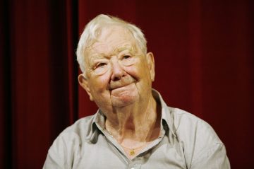 Valkohiuksinen ja vaaleanharmaaseen paitaan pukeutunut iäkäs mies hymyilee lempeästi istuen syvänpunaisen verhon taustalla.