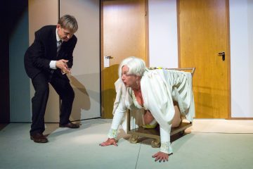 Mustaan pukuun pukeutunut mies seisoo ja osoittaa elein kohti valkoisiin hiuksiin pukeutunutta vanhempaa valkoista naista, joka ryömii nelinkontin pyöräkärryn päällä huoneessa, jossa on puuovet.