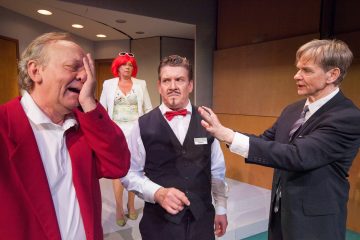 Neljä näyttelijää näyttämöllä: punatakkinen mies pitelee päätään hädissään, punatukkainen aurinkolasipäinen nainen seisoo hänen takanaan, liivipäinen mies nyrpistelee otsaansa, ja toinen pukumies viittoo kohti ryhmää.