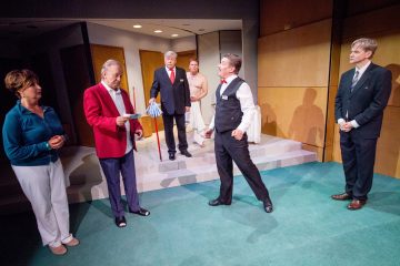 Kuusi näyttelijää esiintyy kirkkaasti valaistulla näyttämöllä. Yksi punatakkinen mies pitelee papereita, kun taas toinen liiviin pukeutunut mies elehtii eloisasti. Muut katselevat tarkkaavaisesti. Taustalla on portaita, ovia ja beigejä seiniä.