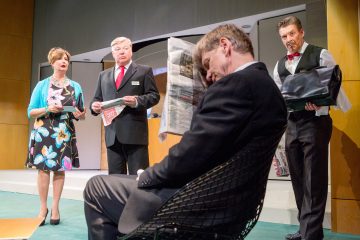 Lavalla on neljä aikuista juhlavaatteissa; yksi nainen ja kaksi miestä seisovat, kun taas toinen mies istuu ja lukee sanomalehteä. Oikealla seisova mies pitää salkkua. Kaikki näyttävät olevan mukana dramaattisessa kohtauksessa.