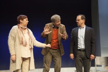 Lavalla seisoo kolme aikuista keskustelemassa. Vasemmalla oleva nainen elehtii ja katsoo keskellä olevaa miestä, jolla on kädet koholla ja eloisa ilme. Oikeanpuoleinen mies katsoo heitä ja vaikuttaa vakavalta.