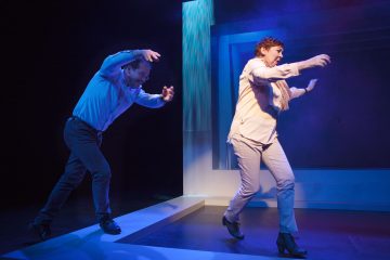 Kaksi ihmistä näyttämöllä, kädet kohotettuna ja taivutettuina, näyttävät näyttelevän dramaattisesti sinisen ja violetin valaistuksen alla, ja heidän takanaan on abstrakteja lavasteita ja heidän liikkeitään korostaa valonheitin.