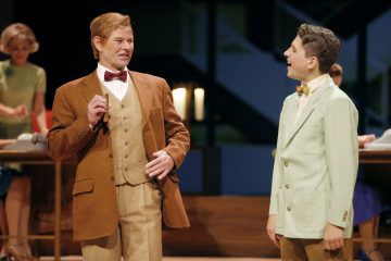 Lavalla seisoo kaksi vanhaan pukuun ja rusettisolmioon pukeutunutta miestä, jotka näyttävät keskustelevan ja hymyilevän. Toisella on ruskea puku, toisella vaaleanvihreä takki. Taustalla kaksi naista työskentelee työpöydän ääressä lämpimässä näyttämövalaistuksessa.