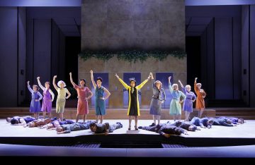 Ryhmä värikkäisiin mekkoihin pukeutuneita naisia seisoo rivissä nyrkkejä kohottaen, kun taas toinen nainen seisoo keskellä kädet ylhäällä. Useita ihmisiä makaa lattialla heidän edessään kirkkaasti valaistulla näyttämöllä.