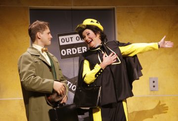 Vihreään pukuun ja rusettisolmioon pukeutunut mies seisoo iloisen mustakeltaiseen pukuun pukeutuneen naisen vieressä. Nainen viittoo leveästi hymyillen oven eteen, jonka keltaisella seinällä on Out of Order -kyltti.