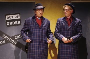 Kaksi miestä, joilla on samanlaiset ruudulliset takit, hatut ja punaiset huivit, seisoo vakavana kielletty -kyltin ja varoitusnauhan edessä. Toinen säätää takkinsa, kun taas toinen pitelee pientä esinettä ja katsoo sivulle.