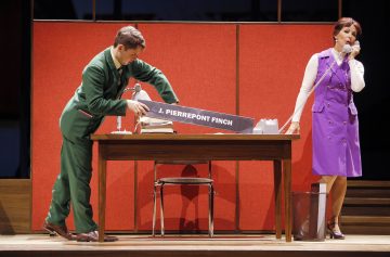 Vihreään pukuun pukeutunut mies asettaa pöydälle nimikyltin, jossa lukee J. Pierrepont Finch, kun taas violettiin mekkoon pukeutunut nainen seisoo vieressä ja puhuu puhelimeen toimistossa, jossa on punaiset väliseinät.