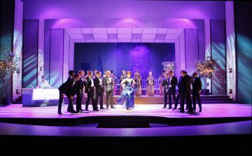 Ryhmä ihmisiä esiintyy kirkkaasti valaistulla lavalla, jossa on violetti valaistus, moderni sisustus ja kaupunkitausta; jotkut seisovat rivissä, kun taas toiset kerääntyvät etualalle.