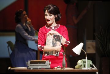 Punapukuinen nainen seisoo pöydän takana hymyillen ja Vogue-lehti kädessään. Pöydällä on kirjoituskone, lamppu ja vihreä kiertävä puhelin. Taustalla näkyy epäselvästi kaksi naista.