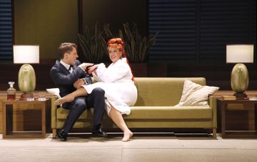 Pukumies ja punatukkainen nainen valkoisessa aamutakissa istuvat vihreällä sohvalla leikkisästi vuorovaikutuksessa. Sarjaan kuuluu kaksi sivupöytää, joissa on vihreät lamput, tyynyjä ja kasvi taustalla.