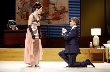 Pukumies polvistuu lattialle, kurottaa kätensä yhteen ja katsoo eleganttiin pukuun ja pitkiin hanskoihin pukeutunutta naista ikään kuin anoen. He ovat lavalla, jonka taustalla on moderneja huonekaluja ja abstraktia taidetta.